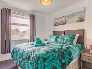 A bedroom with a bed, a window with curtains, a painting on the wall, and a bedside table with a lamp.
