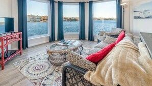 Een gezellige woonkamer met een groot raam met uitzicht op een waterpartij, een televisie op een standaard en een comfortabele zithoek met een kamerkleed.