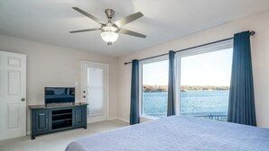 Een slaapkamer met een bed, een televisie op een kast, een plafondventilator en een groot raam met uitzicht op een wateroppervlak.