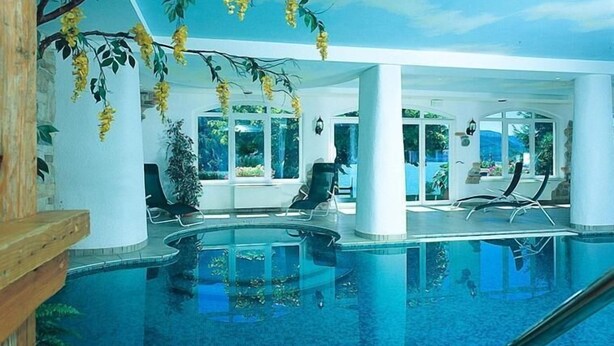 A large indoor swimming pool with clear blue water, surrounded by white pillars and large windows. - Hotel Des Alpes (Molveno)