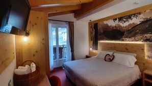 A bedroom with a bed, a television, a wooden headboard, and a window with a mountain view. - Hotel Des Alpes (Molveno)