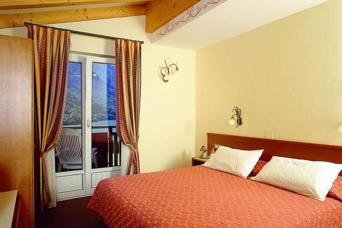 Ein Hotelzimmer mit Bett, Vorhängen, einem Nachttisch, einer Wandlampe und Blick durch eine Tür.