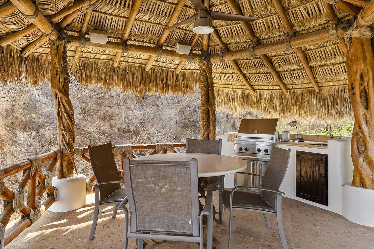 Un espace extérieur aménagé pour les repas, doté d’un toit de chaume, d’un barbecue et offrant une vue sur le paysage environnant.