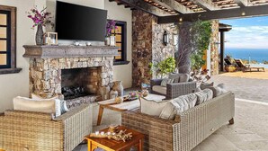 A stone fireplace with a flat-screen TV above it, wicker furniture, and a view of the ocean. - Ultra Private Villa w/ Open Floor Plan, Premium Bedding, & Big Ocean View. Daily Butler Included (San Jose del Cabo)