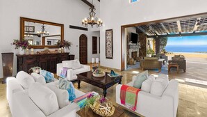 A spacious living room with white sofas, a wooden coffee table, and a chandelier. There is a large mirror on the wall and a view of the beach through an open patio area. - Ultra Private Villa w/ Open Floor Plan, Premium Bedding, & Big Ocean View. Daily Butler Included (San Jose del Cabo)