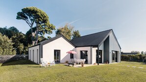 Uma casa moderna de dois andares com um amplo espaço de varanda, conjunto de jantar e uma grande árvore ao fundo.