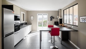 Uma cozinha moderna com geladeira, forno e pia de aço inoxidável, uma área de jantar com uma cadeira vermelha e grandes janelas que proporcionam luz natural.