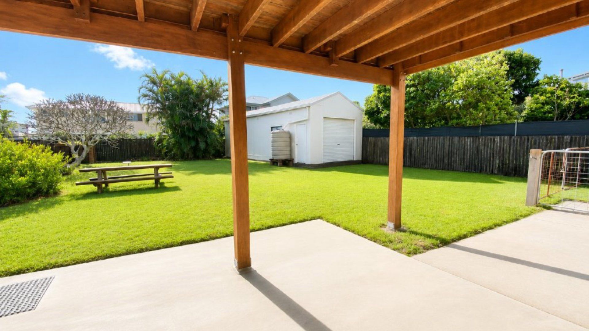 Un patio couvert avec un sol en béton, un banc en bois et une cour arrière enherbée, agrémentée d’un garage blanc et d’une clôture.