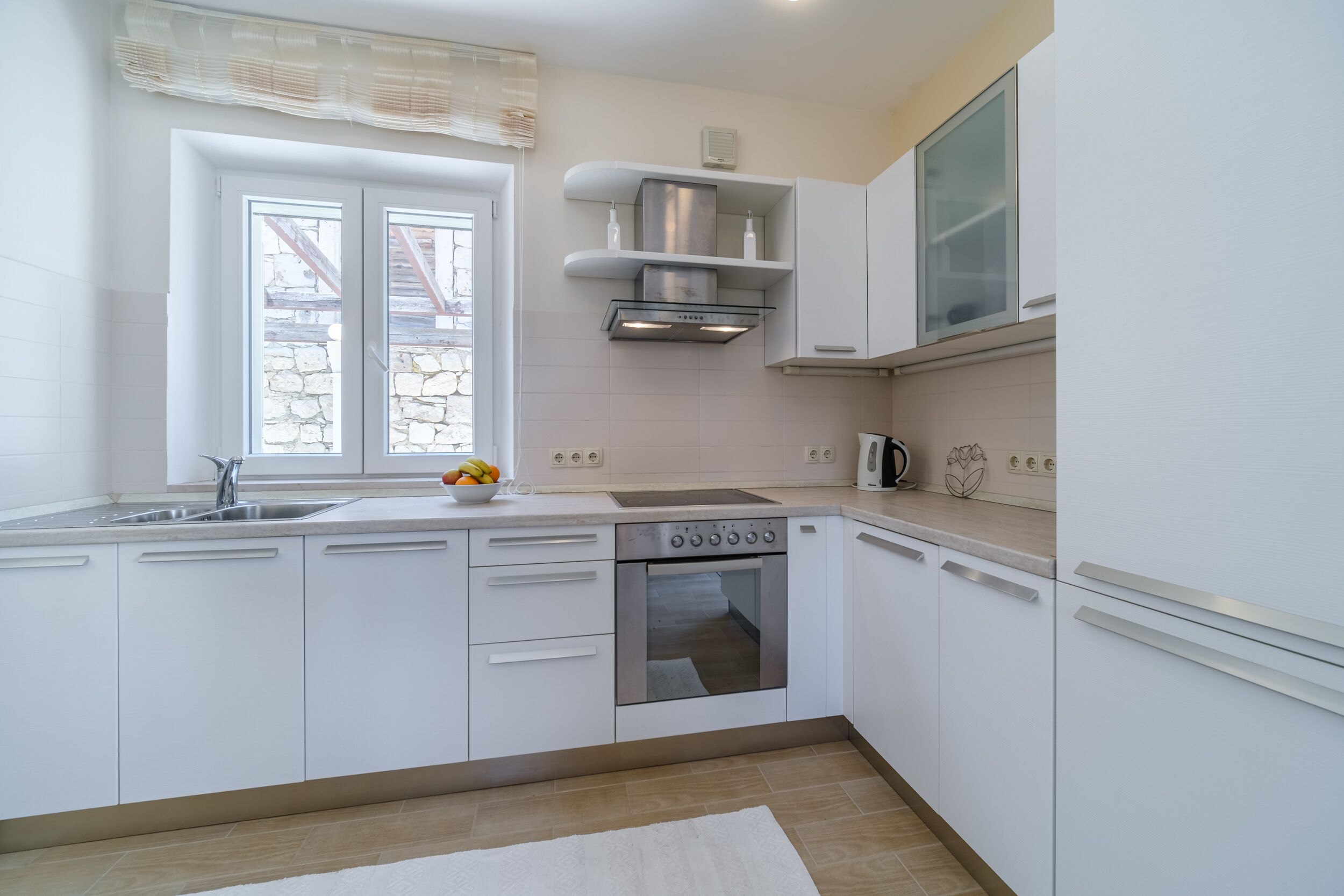 A modern kitchen with white cabinets, a built-in oven, a stainless steel range hood, and a window with a view of a stone wall.