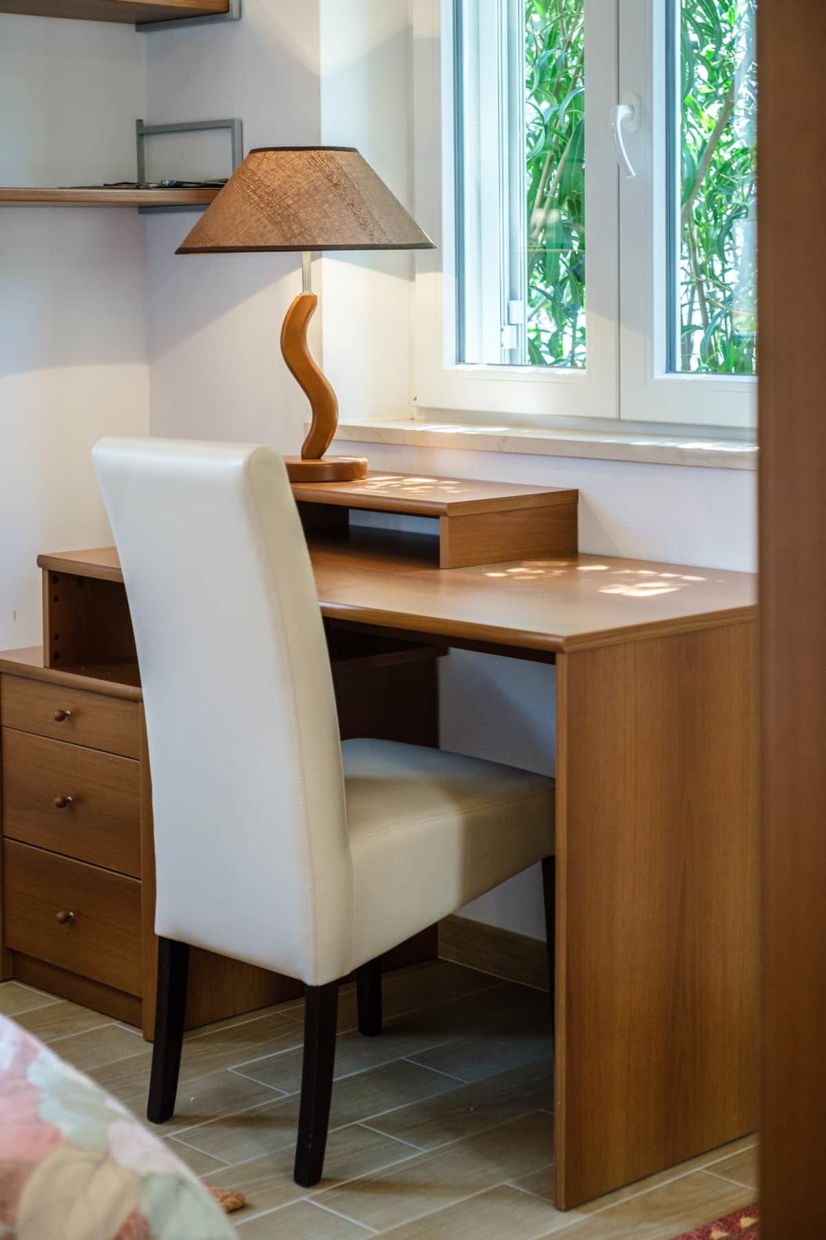 A wooden desk with a white chair, a lamp, and a window with a view of greenery.