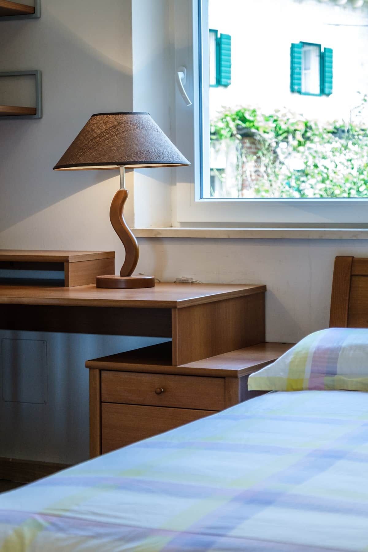 Ein Schlafzimmer mit einem Bett, einem Holztisch, einer Lampe und einem Fenster mit Blick auf Grünflächen.