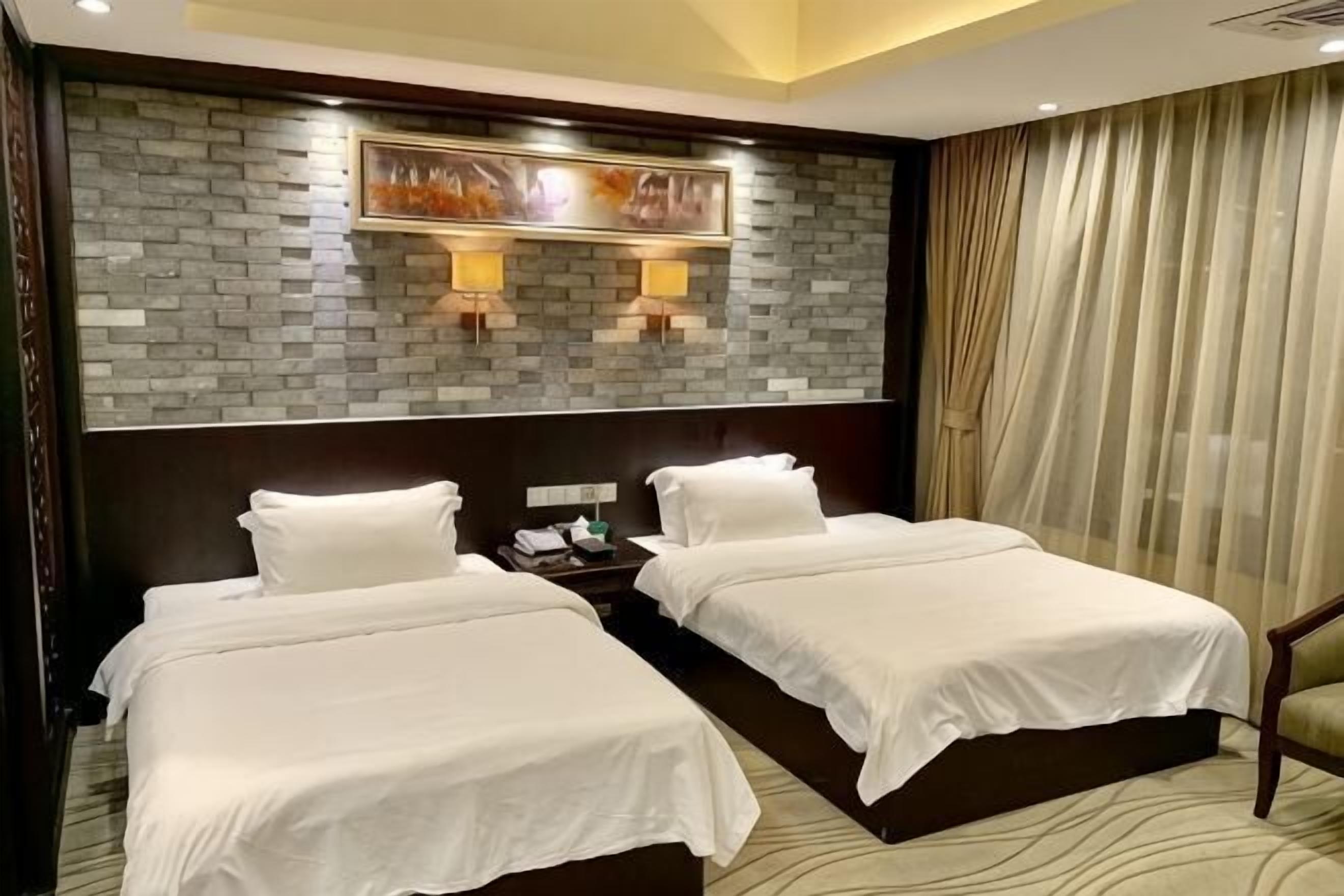 A hotel room with two beds, a brick-patterned wall, two wall-mounted lamps, a nightstand, and a large window with curtains.