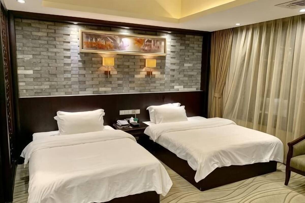 Quarto de hotel com duas camas, parede com padrão de tijolos, duas luminárias fixadas na parede, mesa de cabeceira e uma janela grande com cortinas.