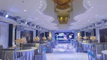 A spacious banquet hall with round tables, chairs, and a stage at the far end.
