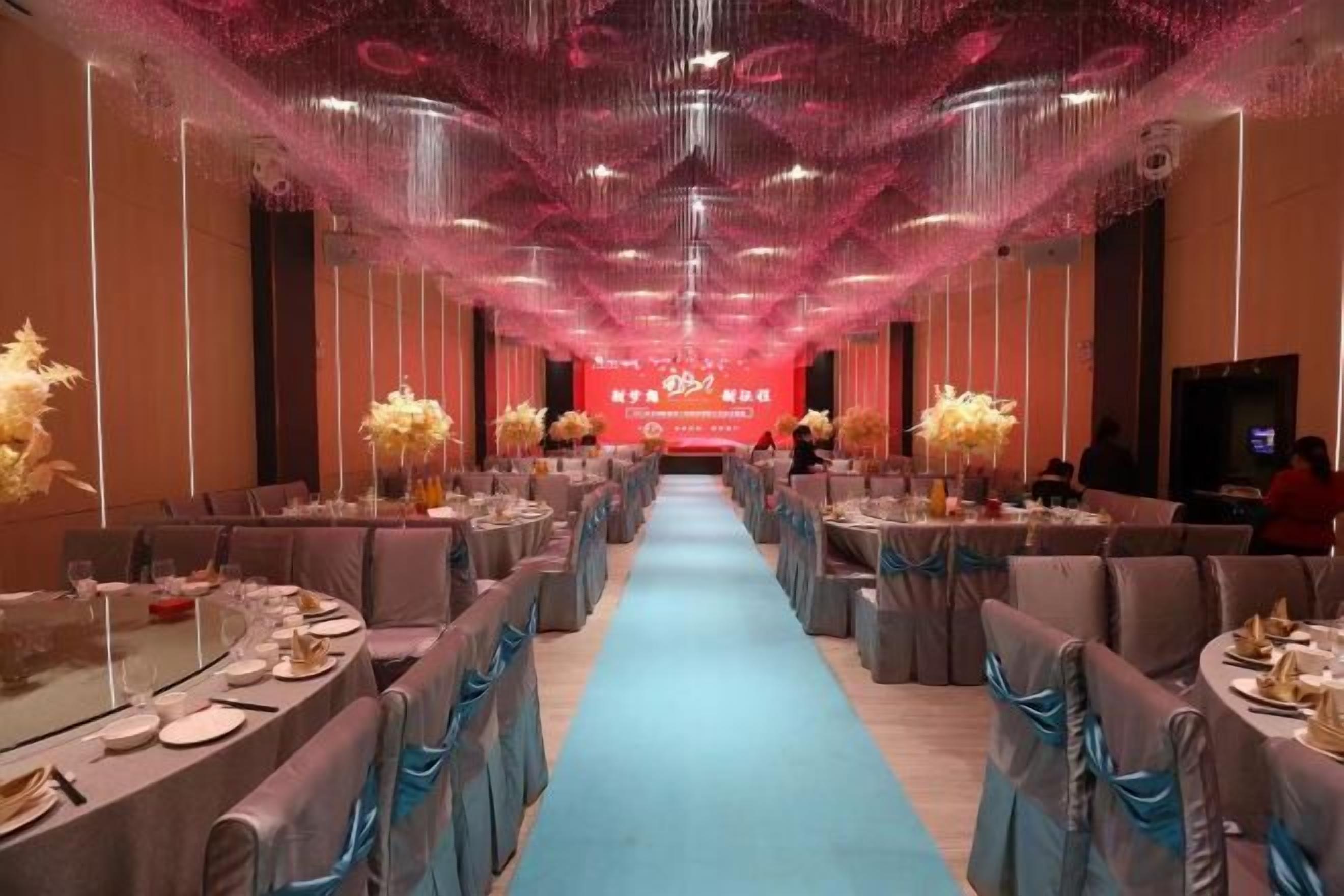 A banquet hall with tables set for a formal event, a stage with a red backdrop, and a blue carpet leading to the stage.