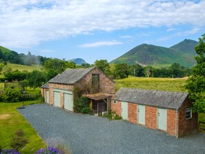 A rustic two-story building with a gravel driveway, surrounded by lush greenery and mountains in the background. - Swinside Lodge (Keswick)