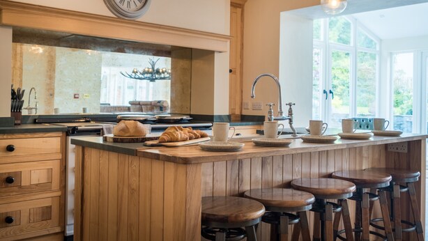 Una cocina con una isla de madera, taburetes de bar, un reloj y una ventana con vista a la vegetación.