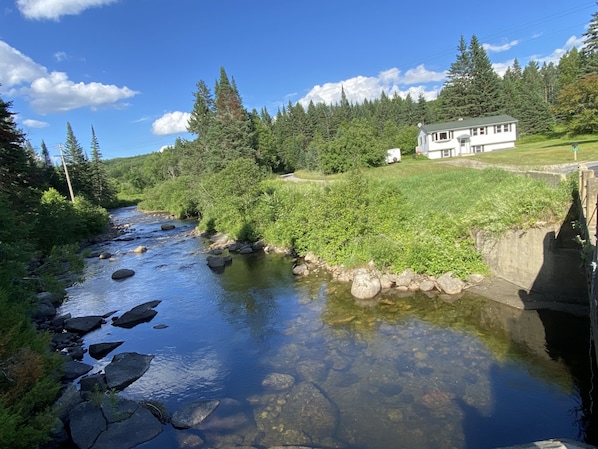 Une maison située au bord d’un ruisseau aux eaux claires, avec des rochers, et entourée d’arbres et de verdure.