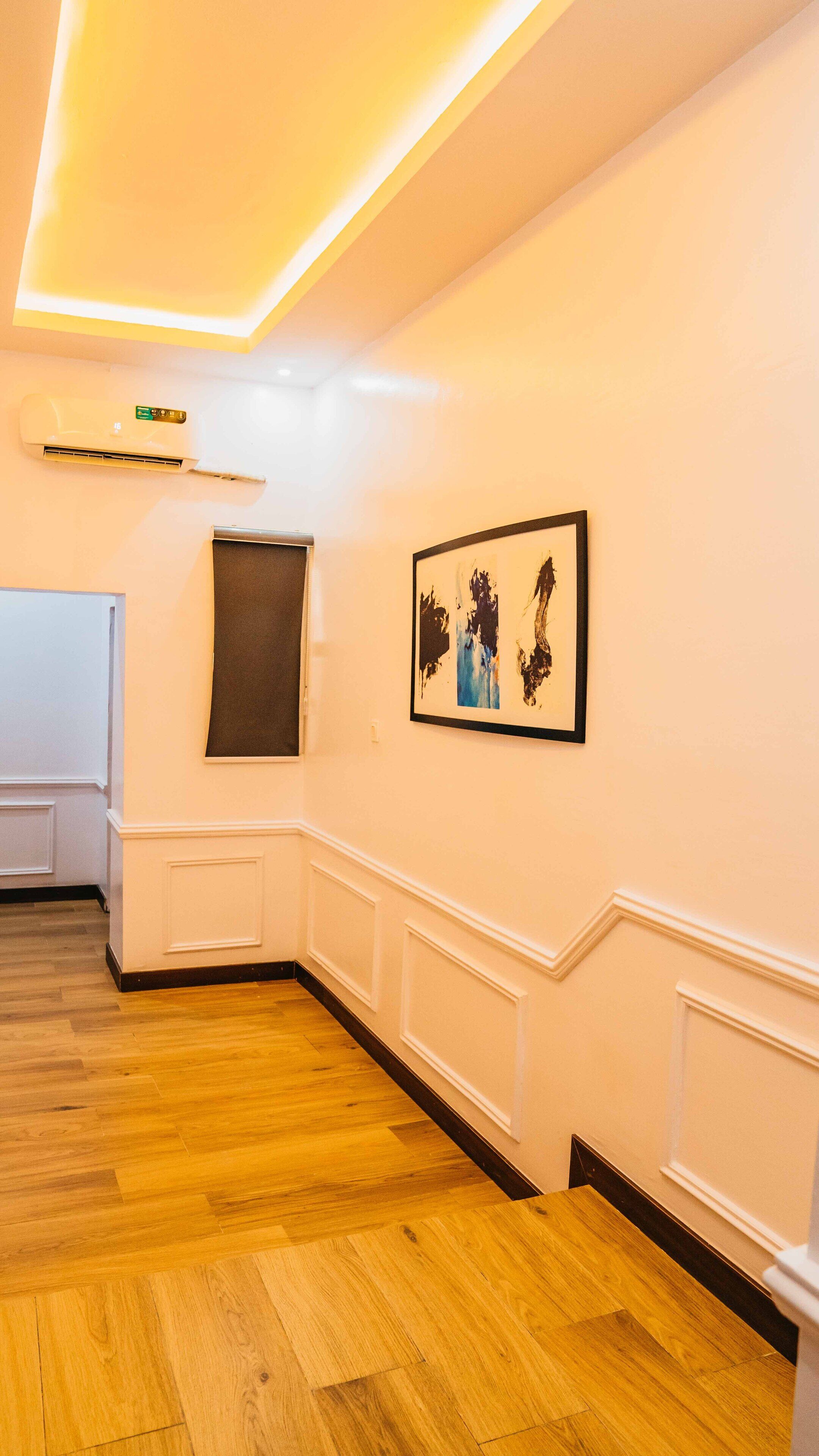 A hotel room with a wooden floor, white walls, a ceiling light fixture, a window with a dark blind, and a framed abstract painting.