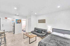 Quarto de hotel moderno com cama, sofá, mesa de centro e área de refeições com cadeiras.