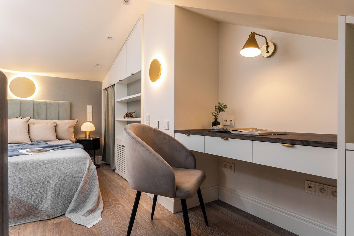 Een moderne slaapkamer met een bed, nachtkastje en een stoel. Er is een bureau met een lamp en een kleine plant. De kamer heeft een neutraal kleurenpalet en houten vloeren.