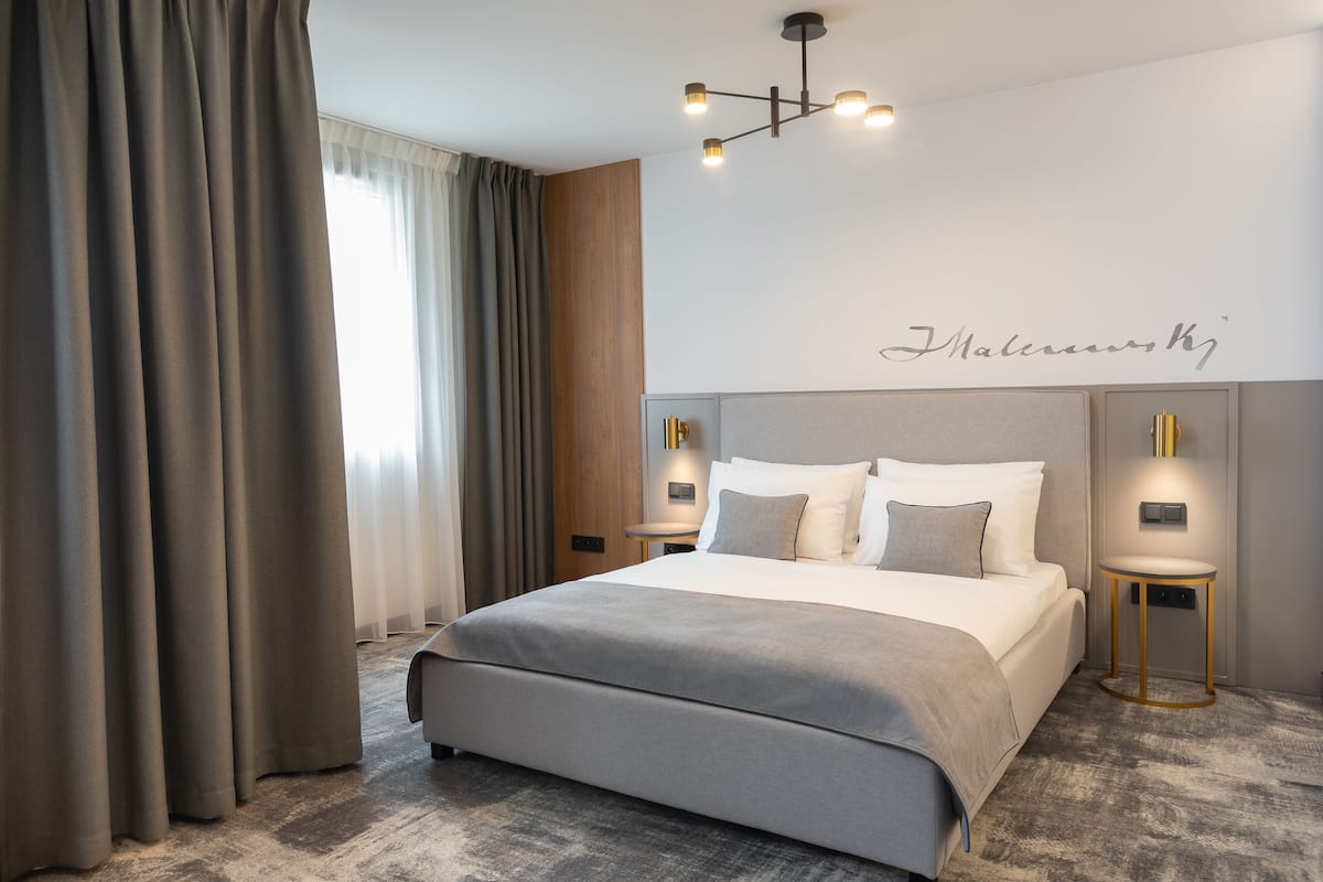 Ein modernes Hotelzimmer mit einem großen Bett, zwei Nachttischen mit Lampen, einer Deckenleuchte und einem großen Fenster mit Vorhängen.