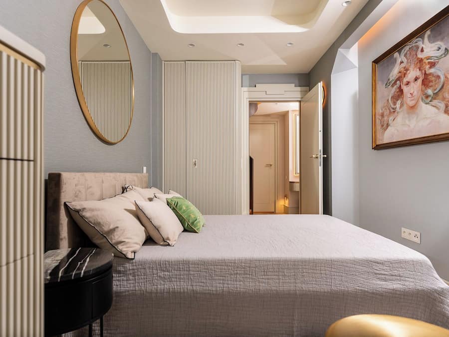 A bedroom with a bed, a round mirror, a painting, and a door leading to another room.