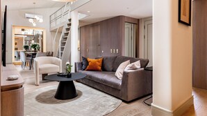 Een moderne woonkamer met een grijze bank, een ronde zwarte salontafel en een witte fauteuil. Er is een trap in de achtergrond te zien, die naar een hoger niveau leidt.