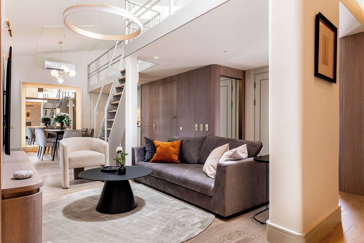 Ein modernes Wohnzimmer mit einem grauen Sofa, einem runden schwarzen Couchtisch und einem weißen Sessel. Im Hintergrund ist eine Treppe zu einer oberen Ebene zu sehen.