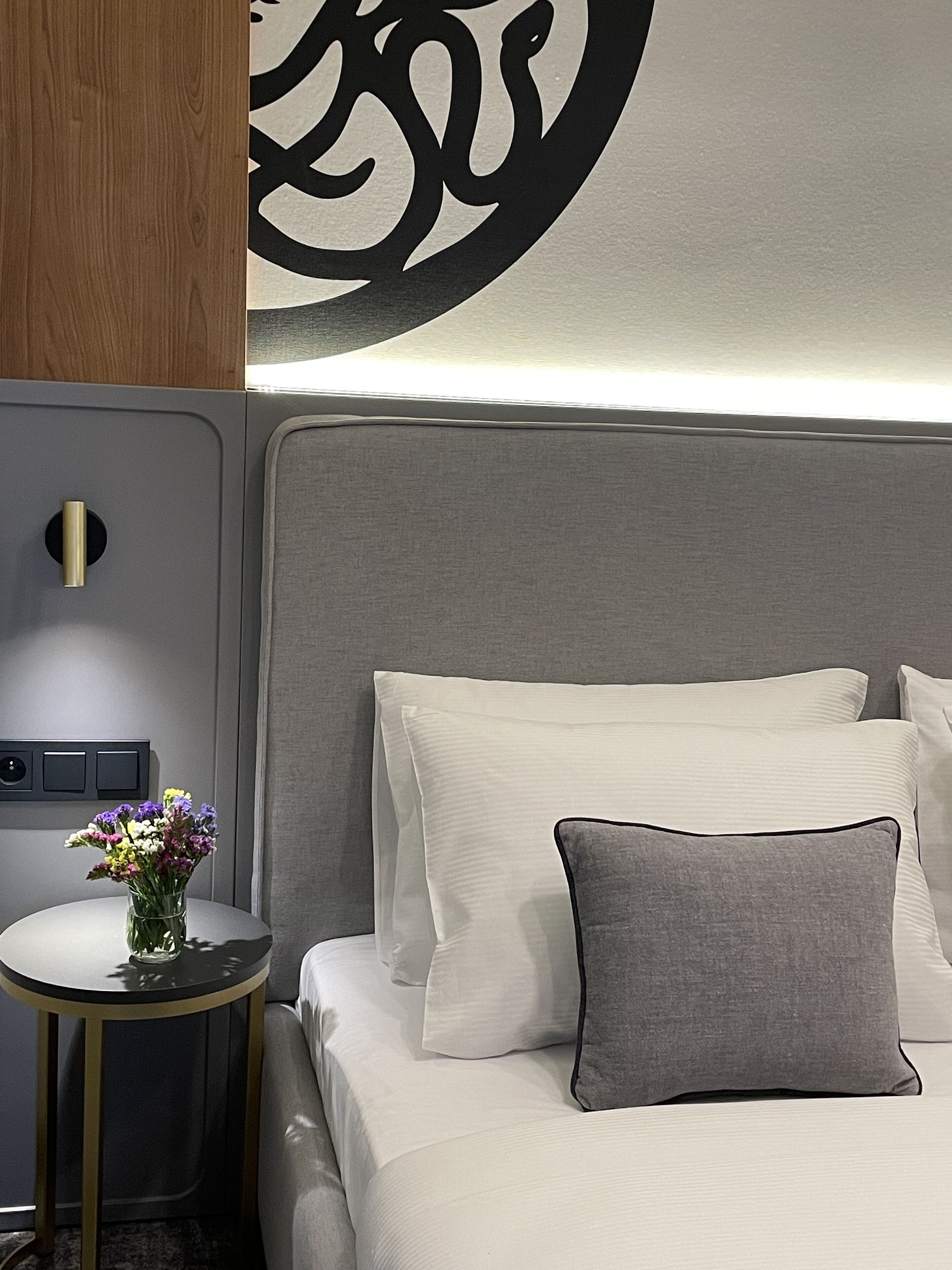 A neatly made bed with white and gray bedding, a small round table with a vase of flowers, and a wall-mounted light fixture.