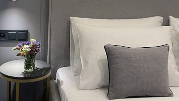 Un lit bien fait, avec une literie blanche et grise, une petite table ronde ornée d’un vase de fleurs et un luminaire fixé au mur.