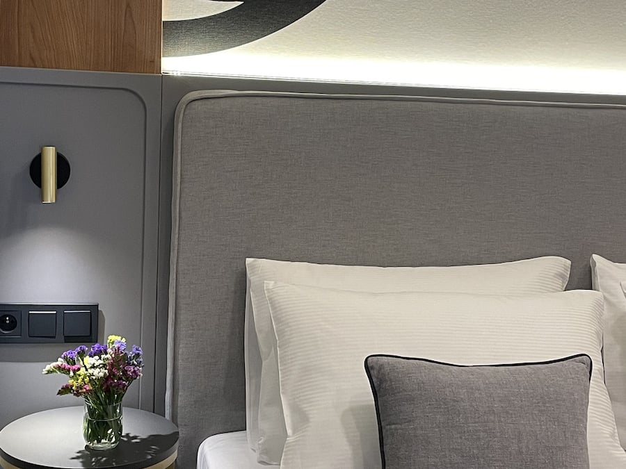 Un lit bien fait, avec une literie blanche et grise, une petite table ronde ornée d’un vase de fleurs et un luminaire fixé au mur.
