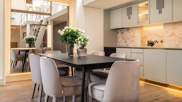 Een moderne keuken met grijze kastjes, een marmeren achterwand en een eethoek met een donkere tafel en grijze stoelen.