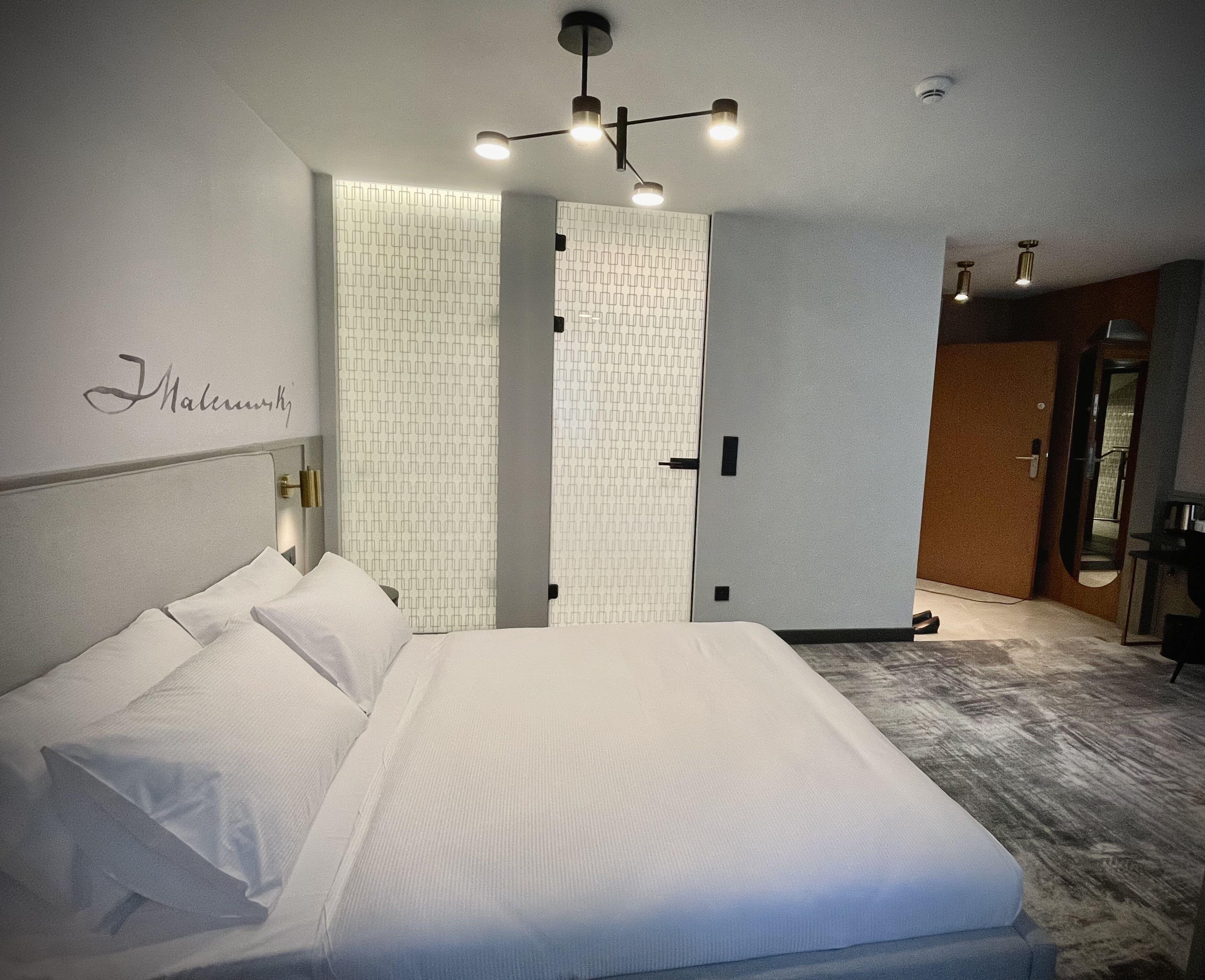 A modern hotel room with a large bed, a headboard with a nameplate, a patterned wall, and a ceiling light fixture.