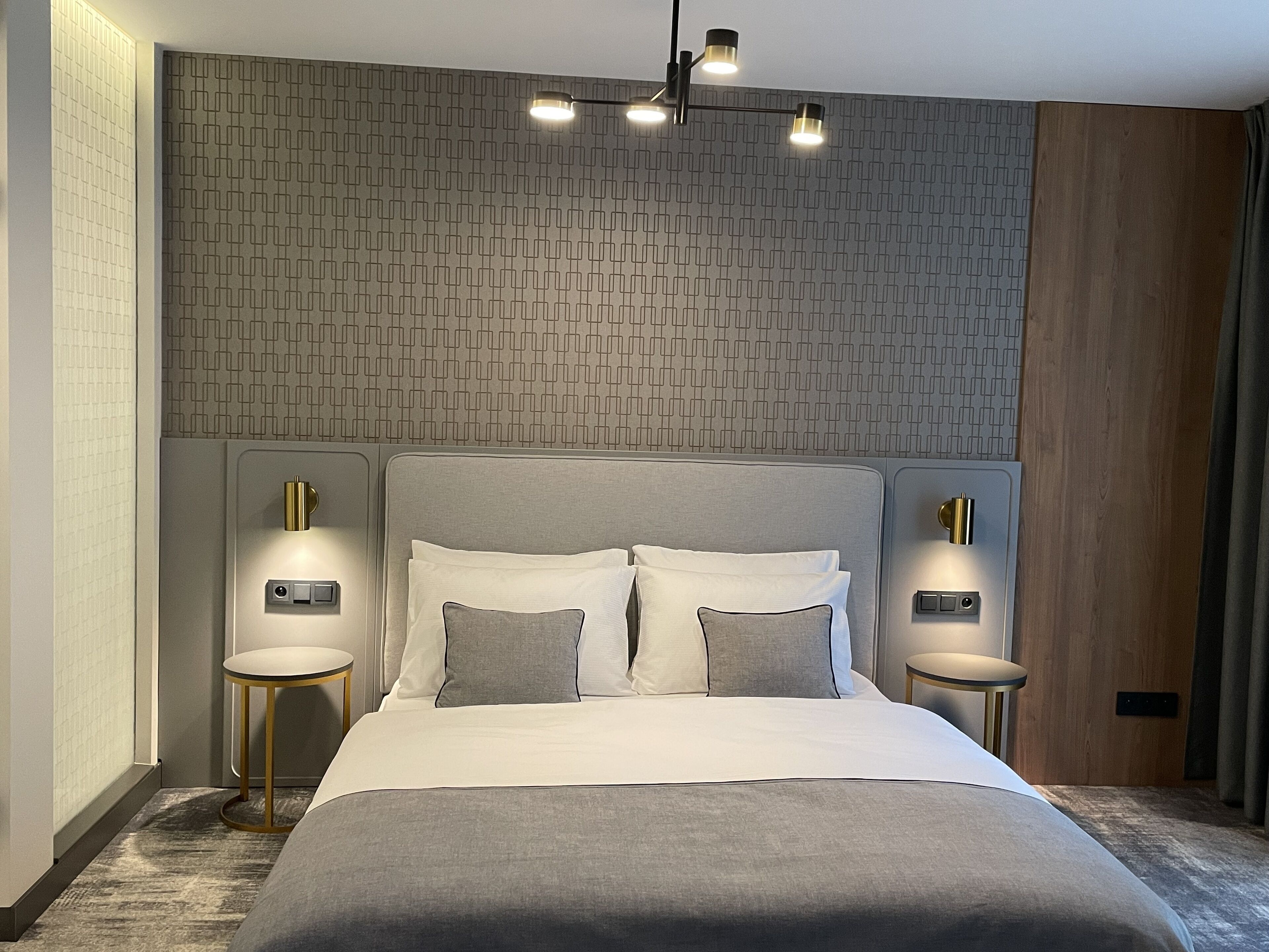 A modern hotel room with a large bed, two bedside tables, a patterned wall, and a ceiling light fixture.