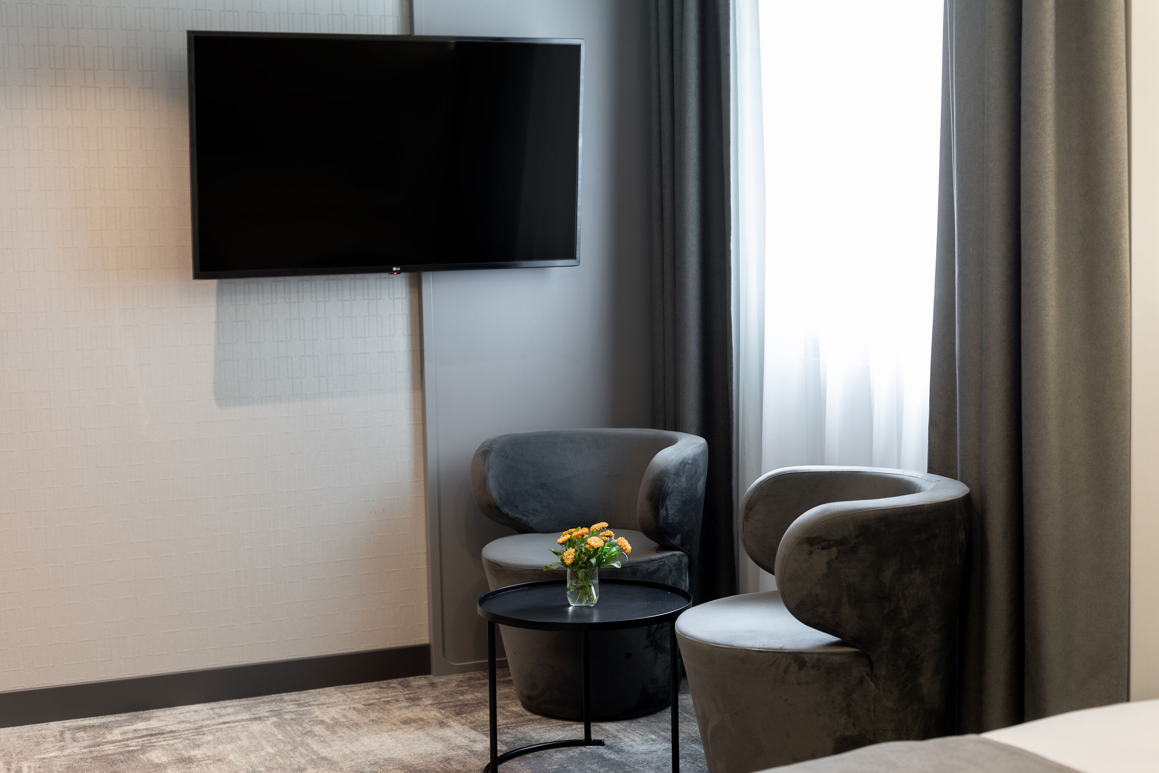 Quarto de hotel moderno com televisão de tela plana na parede, uma pequena mesa redonda com um vaso de flores e duas poltronas cinzas.