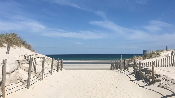 Ein sandiger Strandweg führt zum Meer, begleitet von Holzzaun und spärlicher Vegetation.