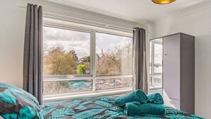 A bedroom with a large window, a bed with a tropical leaf pattern bedspread, a grey wardrobe, and a hanging light fixture. - Lovely Apartment 3 King-size Beds (Torquay)