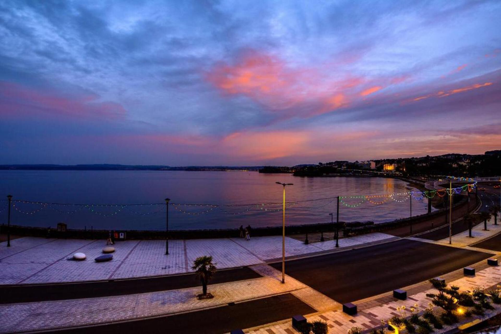 A waterfront promenade with a paved walkway, street lamps, and a view of the sea at dusk.