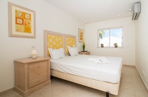Une chambre à coucher comprenant un lit, des tables de chevet, une commode, une fenêtre avec des plantes et des œuvres d’art accrochées aux murs.