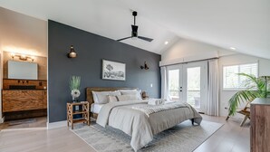 A bedroom with a large bed, a ceiling fan, a bedside table with a lamp, a mirror, and a window with curtains.