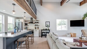 A modern kitchen with a central island, wooden stools, and a dining area with a sofa and TV.