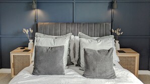 A bedroom with a grey upholstered headboard, two bedside tables with lamps, and a bed with grey and white pillows.