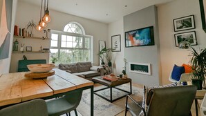 A spacious living room with a large window, a wooden dining table, a sofa, and a coffee table. There are shelves with books and decorative items, and a TV mounted on the wall.