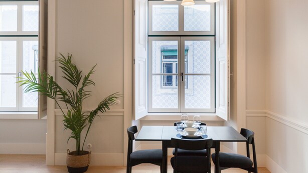 A dining area with a table set for two, a potted plant, and a window with a view of a building. - FLH Praça Rossio Elegant Flat (Lisbon)