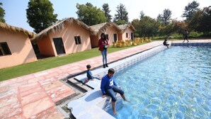 Piscina com deck de pedra, rodeada por cabanas com telhados de palha e uma área gramada.