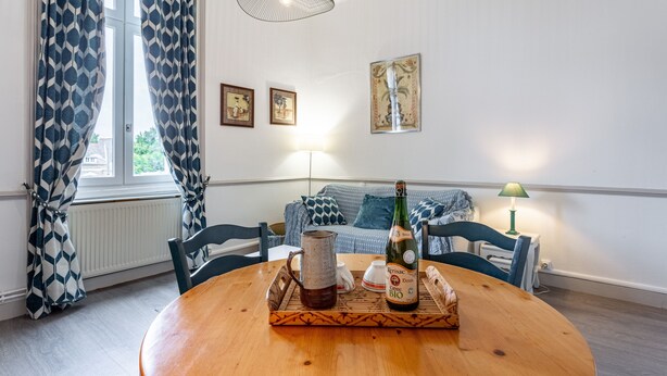 Een gezellig eetgedeelte met een houten tafel, een fles wijn en een karaf. Er staan twee stoelen, een bank en een lamp op de achtergrond.