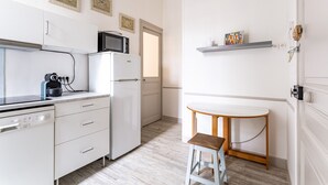 Een kleine keuken met witte kastjes, een magnetron, een koelkast en een eethoekje met een houten tafel en krukje.