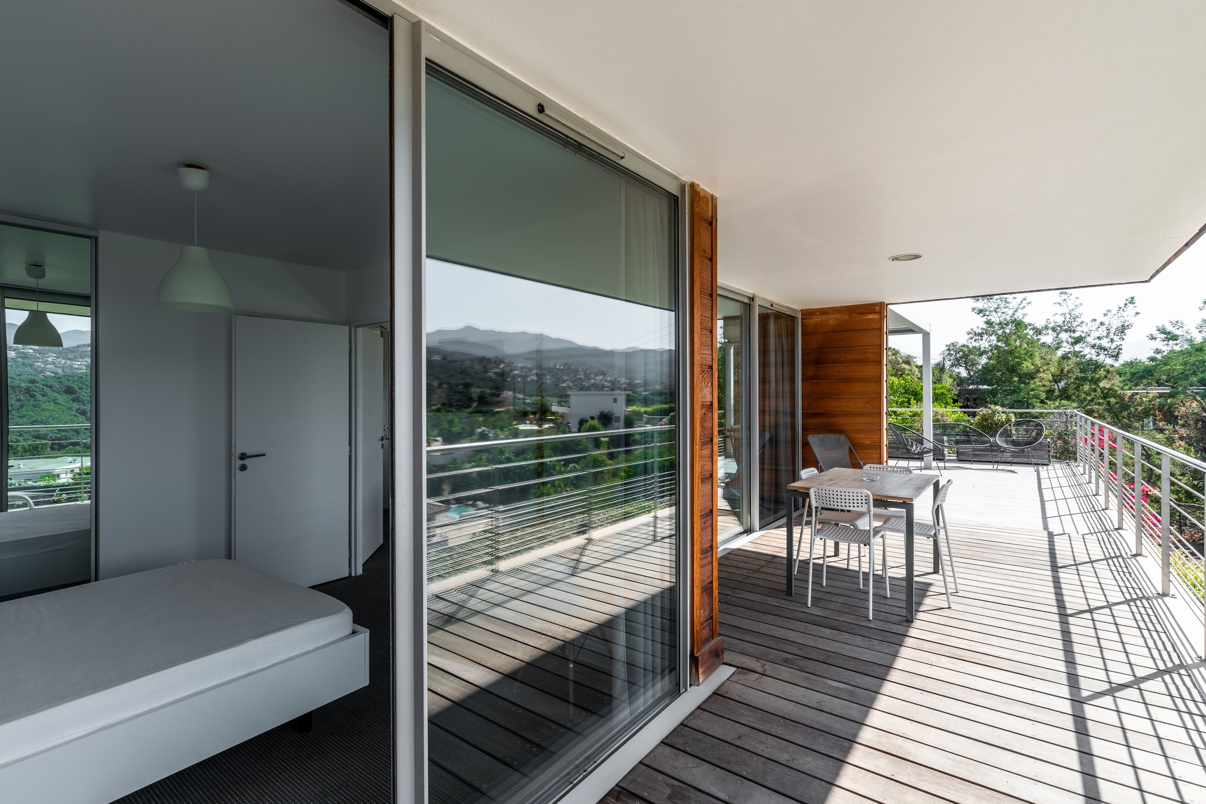 Un balcon moderne avec une terrasse en bois, une table et des chaises blanches, et une vue sur le paysage urbain et les montagnes.
