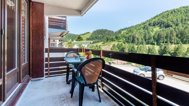 Un balcon avec une table et des chaises, donnant sur une route et des collines verdoyantes.